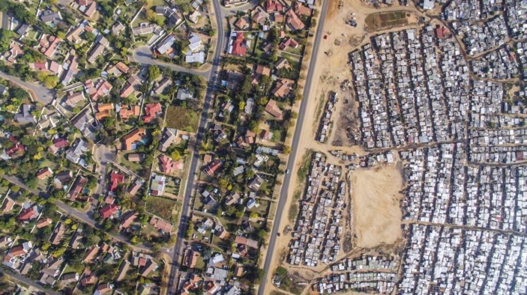 Aerial view showing a stark divide between two neighboring areas in South Africa: on the left, a suburb with spacious homes, tree-lined streets, and green yards; on the right, a densely packed informal settlement with tightly clustered small structures and unpaved ground. A single road runs between the two contrasting communities.