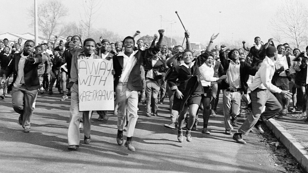 Black and white photo of a large group of young protesters marching down a street, many with raised fists and expressive faces. One person at the front holds a sign reading "AWAY WITH AFRIKAANS," indicating opposition to the use of Afrikaans, likely in an educational or political context. The image captures a moment of collective resistance during South Africa's apartheid era.
