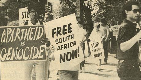 A group of protesters march outdoors holding large signs that read messages such as “APARTHEID IS GENOCIDE,” “FREE SOUTH AFRICA NOW!,” and “STOP APARTHEID NOW!” The black-and-white image captures people walking together in a demonstration against apartheid.