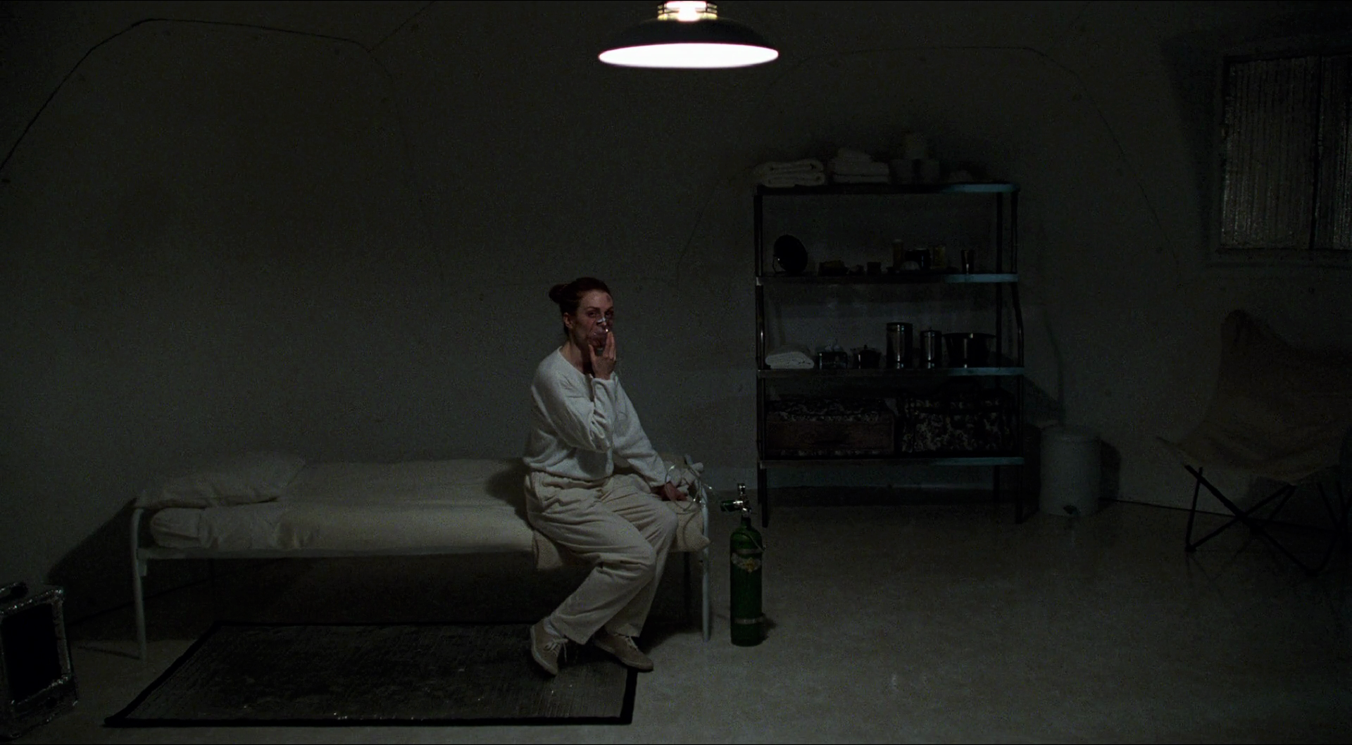 A person dressed in light-colored clothing sits on the edge of a narrow bed inside a dim, concrete, bunker-like room. They hold an oxygen mask to their face, connected to a green oxygen tank on the floor. A single overhead lamp casts harsh light, and metal shelves with jars, containers, and folded linens line the wall, creating a stark, claustrophobic atmosphere.