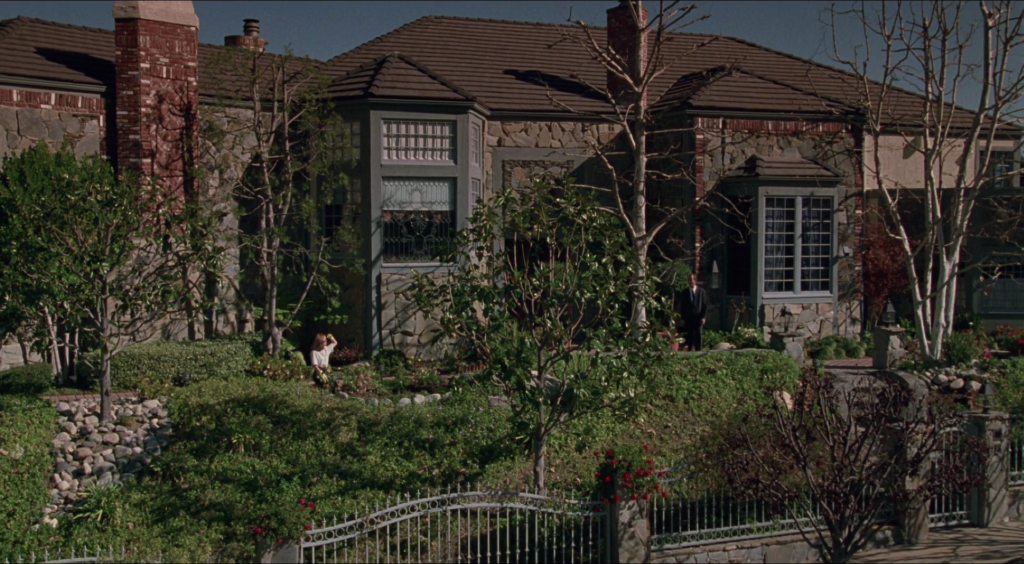 A wide shot of a large stone and brick house with a tiled roof, surrounded by dense greenery and landscaped gardens. A woman sits low in the yard near flowers, while a man in a dark suit stands near the front entrance. Leafless trees frame the scene, and a metal fence runs along the front of the property.