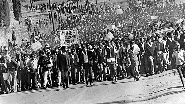 A crowd of Black students marching in the street during the Soweto Uprising, many holding signs and raising their fists, with a few police or military vehicles visible in the background.